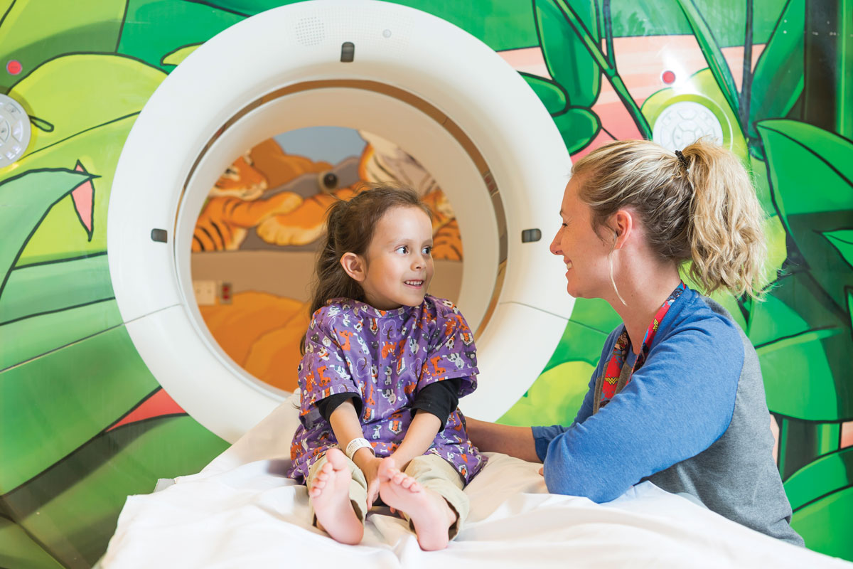 Girl sitting on CAT scan machine smiling at nurse next to bed