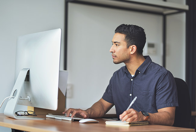 Man sitting at desk on computer, typing and taking notes in a notebook