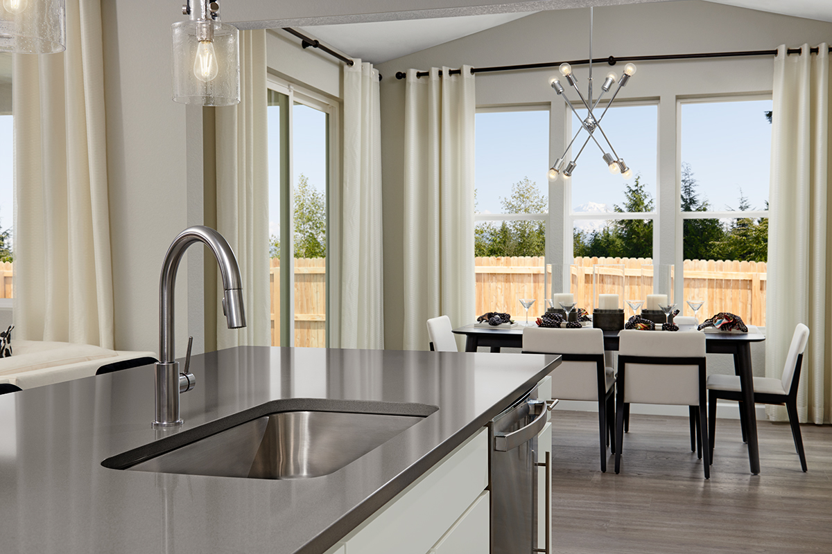 Island with gray countertop in front of dining room with sliding doors and three tall windows with tall curtains