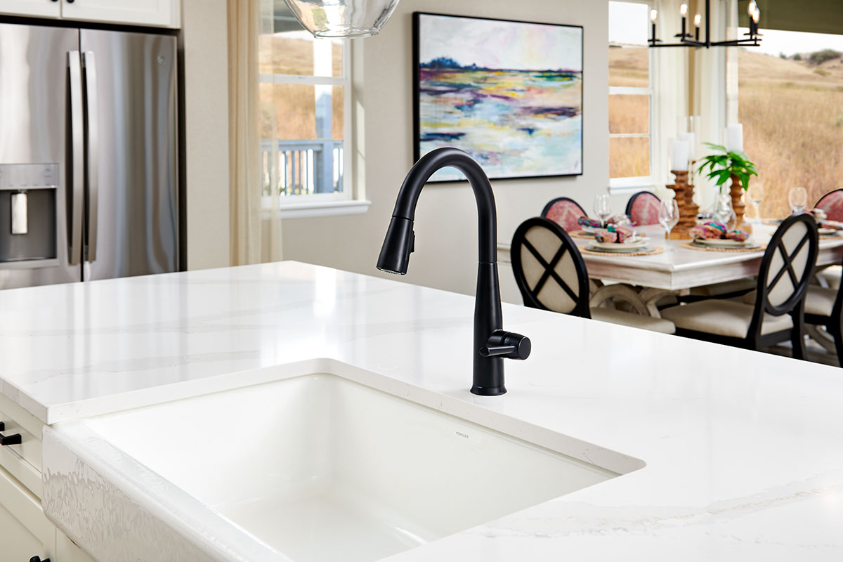 Farmhouse kitchen sink on island with black faucet and refrigerator and dining table in background